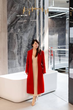 Modern bir küvetin yanındaki lüks banyo galerisinde yürüyen, kırmızı ceketli ve bej elbiseli bir kadın mimar.