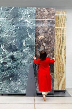 Kırmızı ceketli bir kadın, bir yapı malzemesi deposunda mermer levhalar seçiyor, gelecek bir tasarım projesi için seçeneklere odaklanıyor.