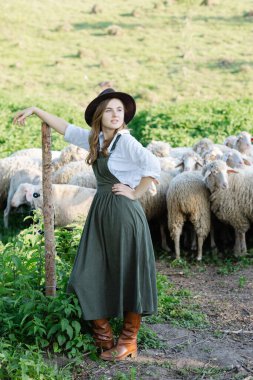 Şapkalı ve tulumlu genç çoban kız bir tahta direğe yaslanmış bir tarladaki koyun sürüsünü izliyor.