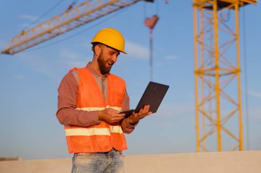 İnşaat mühendisi arkaplanda vinç olan inşaat alanında dizüstü bilgisayar kullanıyor, projeyi yönetiyor ve güvenliği sağlıyor