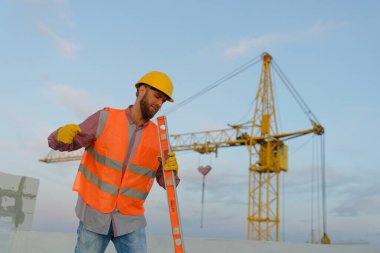 İnşaat işçisi yüksek görünürlük yeleği giyiyor ve arka planda vinç ile inşaat sahasında ruh seviyesini koruyor.