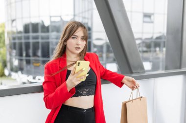 Cheerful young woman dressed in stylish suit holding mobile phone in hand outdoors. Beautiful girl holding mobile and standing on street with paper bags