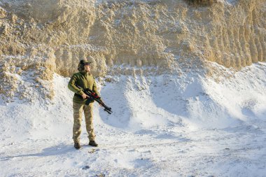 Karlı bir arazide elinde tüfekle duran ve kamuflaj giysileri giyen bir asker kış koşullarında askeri varlığını sergiliyor.