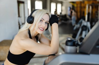 Sporcu kadın koşu bandına yaslanırken gülümsüyor, modern bir spor salonunda antrenmanının tadını çıkarıyor, vücut bulmuş spor yapıyor ve sağlıklı bir hayat sürüyor.