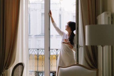 Adult woman enjoying a relaxing morning holding a cup of coffee or tea on a Parisian apartment balcony, smiling at the camera