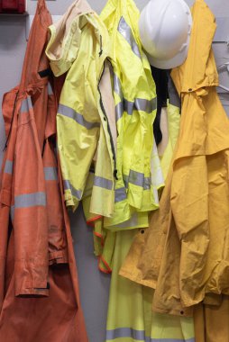 Turuncu ve sarı tonlarda, yüksek görüş mesafesi olan yağmurluklar ve duvarda asılı kasklar. Yansıtıcı çizgiler. 