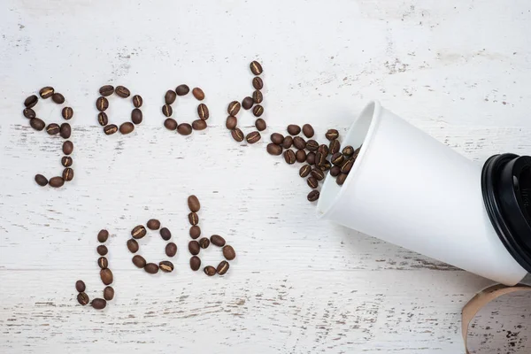 A good job coffee. Message of good job spelled out of coffee beans spilling out of a white paper cup onto a white-washed wooden plank background.