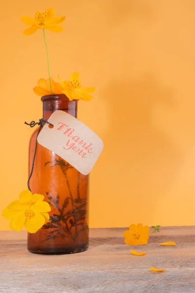 Thank you note tag on an antique amber glass bottle holding yellow flowers.  Minimalist.  yellow background  and wooden table.