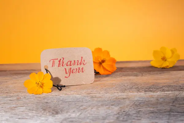Thank you note on recycled paper tag on a wooden background with bright yellow-orange flowers.  Minimalistic.