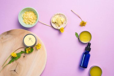 Karahindiba merhemi, karahindiba yağı, balmumu, çiçekler ve yapraklar beyazlatılmış tepside, mor arka plan.