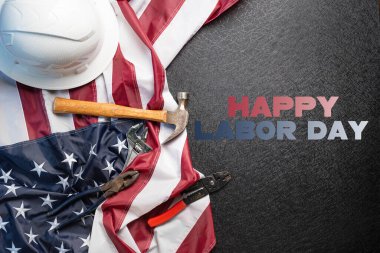 Çalışma araçları ve güvenlik ekipmanlarıyla siyah zemin üzerinde Amerikan bayrağı Mutlu İşçi Bayramı metni.