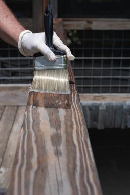 Eldivenli bir el siyah bir boya fırçası tutuyor, ahşap bir güverteye su yalıtıcısı takıyor..