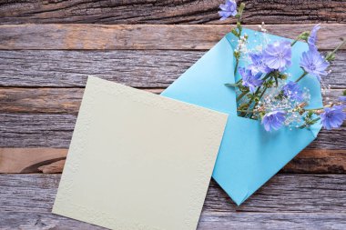 Açık yeşil, boş bir kart, parlak mavi bir zarfın yanında küçük mor çiçeklerle doldurulmuş ahşap bir tepside..