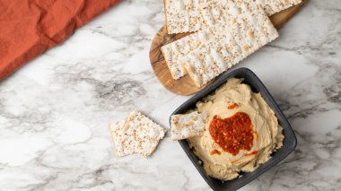 Siyah bir kasede kavrulmuş kırmızı biber humusu. İçinde ot, tuzlu kraker, tahta tahtada yassı ekmek, mermer arka planda turuncu peçete..