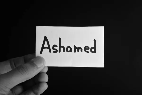 Üzerinde ASHAMED yazan bir kart tutuyor.