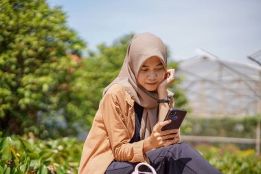 Modern tesettür takan Asyalı kadın parkta akıllı telefonunu kullanarak sosyal medya oynuyor. Akıllı telefonlar pratik olarak kullanılabilen modern teknolojilerdir.
