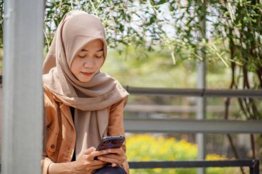 Modern tesettür takan Asyalı kadın parkta akıllı telefonunu kullanarak sosyal medya oynuyor. Akıllı telefonlar pratik olarak kullanılabilen modern teknolojilerdir.