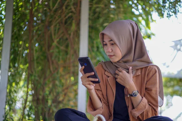 身穿现代头巾的亚洲女人在公园里用她的智能手机玩社交媒体 智能手机是可实际使用的现代技术 — 图库照片