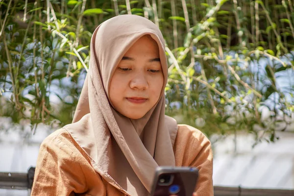 園内には現代のヒジャーブを着たアジア人女性がスマホを使ってSnsをしている スマートフォンは実際に使用できる現代的な技術です — ストック写真