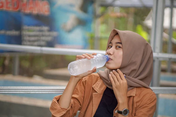 stock image a beautiful muslim asian woman wearing a modern hijab is drinking bottled water in the park