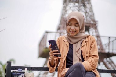 Modern tesettürlü Asyalı bir kadın parkta akıllı telefonunu ve Eyfel kulesinin arka planını kullanarak sosyal medyada oyun oynuyor. Akıllı telefon, pratik olarak kullanılabilen modern bir teknoloji.