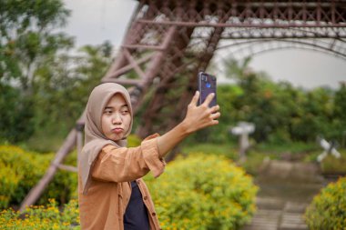 Modern tesettürlü Asyalı kadın akıllı telefonunu kullanarak çiçek bahçesinde selfie çekiyor. Akıllı telefon, pratik olarak kullanılabilen modern bir teknoloji.