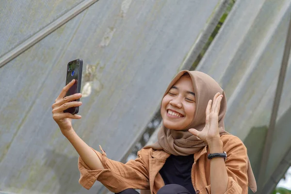 現代のヒジャーブを着たアジアの女性がスマートフォンを使って公園でビデオ通話をしています スマートフォンは実用的に使用できる近代的な技術です — ストック写真