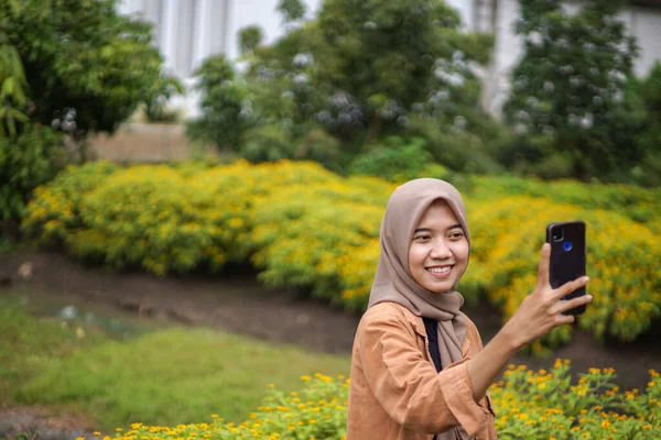 現代のヒジャーブを着たアジア人女性がスマホを使って花畑で自撮りをしている スマートフォンは実用的に使用できる近代的な技術です — ストック写真