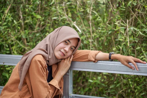 身穿现代头巾服装的美丽的穆斯林亚洲女人坐在不锈钢花园的篱笆上 — 图库照片
