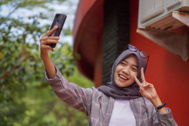 Modern tesettüre bürünmüş güzel Asyalı Müslüman kız öğrenci akıllı telefonunu kullanarak parkta selfie çekiyor.