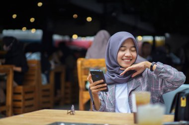 Güzel Asyalı Müslüman kadın modern başörtüsü takıyor gülümsüyor ve bir kafede akıllı telefon tutuyor.