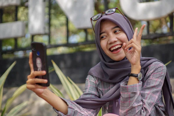 美しいアジア系ムスリム女子大生現代的なヒジャーブを身に着けています彼女のスマートフォンを使用して公園で自殺 — ストック写真