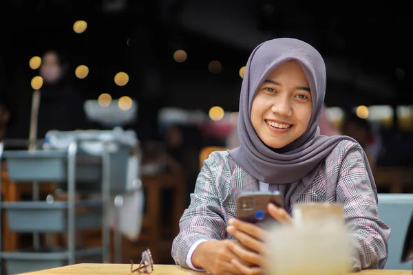 現代のヒジャーブを着た美しいアジア系ムスリム女性が笑顔でカフェでスマホを持ち — ストック写真