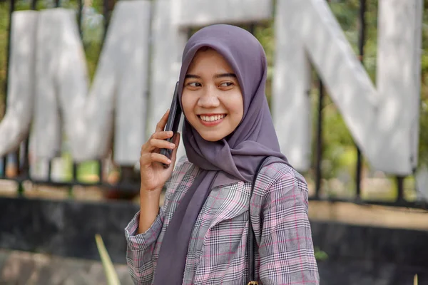現代のヒジャーブを身に着けている美しいアジア系ムスリム女性学生は彼女のスマートフォンを使って公園で誰かを呼んでいます — ストック写真