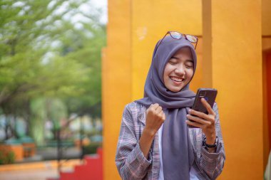 Modern tesettür giyen Asyalı Müslüman kız öğrenci parkta akıllı telefonu tutarken mutlu bir ifadeyle.