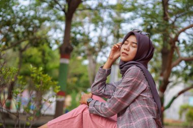 Güzel Asyalı Müslüman kadının modern tesettüre binmiş parkta otururken çekilmiş bir fotoğrafı.