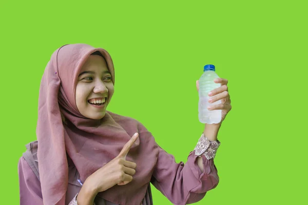 一位漂亮的亚裔穆斯林妇女 头戴头巾 指着手中拿着的水瓶 盖在绿色的背景上 穆斯林妇女戴头巾 手里拿着装有花蜜的瓶装水 — 图库照片