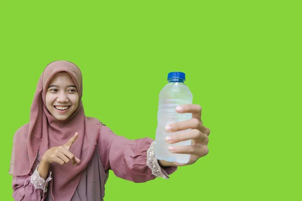 一位漂亮的亚裔穆斯林妇女 头戴头巾 指着手中拿着的水瓶 盖在绿色的背景上 穆斯林妇女戴头巾 手里拿着装有花蜜的瓶装水 — 图库照片