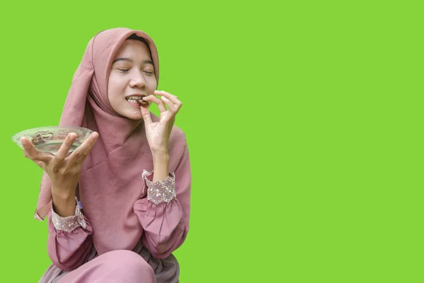 美しいアジア系女性のヒジャーブは緑の背景で断食明けのデートを食べています イスラム教とラマダーンの概念を持つヒジャーブを着たムスリム女性の写真 — ストック写真