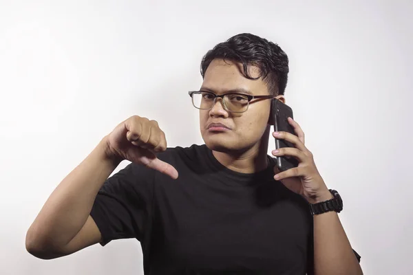 黒のTシャツを着たアジア系の若い男性がスマートフォンで誰かを呼び出し 白い背景に孤立した親指で否定的な表現を示しています — ストック写真