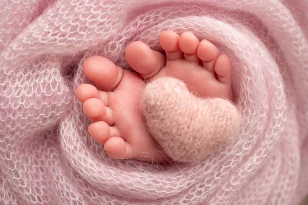 Knitted pink heart in the legs of a baby. Soft feet of a new born in a pink wool blanket. Close-up of toes, heels and feet of a newborn. Macro photography the tiny foot of a newborn baby.