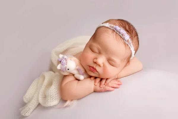 Sleeping newborn girl in the first days of life on a white background. Close-up studio portrait of a child. A newborn baby is wrapped in a white knitted blanket with a white headband.