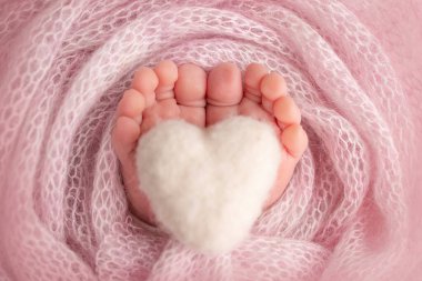 Knitted white heart in the legs of a baby. Soft feet of a new born in a pink wool blanket. Close-up of toes, heels and feet of a newborn. Macro photography the tiny foot of a newborn baby.