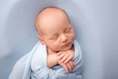 A cute newborn boy in the first days of life sleeps naked on a blue fabric background. The baby is wrapped in a blue winding, a blanket. Studio professional macro photography, portrait of a newborn 