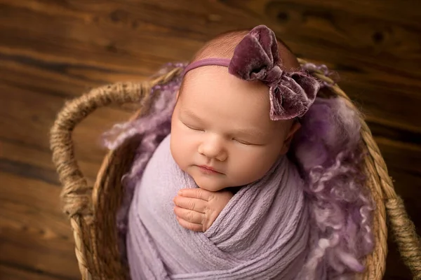 ウィッカーバスケットに天然の茶色の木の背景に生まれたばかりの女の子を眠っています 頭にライラック バイオレットの弓を持つ新生児 紫の背景碑文の愛 新生児の美しい肖像画 — ストック写真