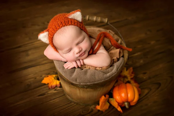 木製のバケツに天然の茶色の木の背景に新生児の女の子を眠っています 彼の頭の上に赤茶色の虎の子のキャップを持つ新生児 赤いカボチャと新生児の美しい肖像画 — ストック写真