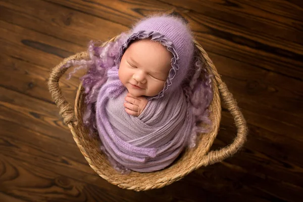 天然の茶色の木を背景に ウィッカーバスケットで新生児の女の子を眠っています 頭にライラック バイオレットキャップを持つ新生児 新生児の美しい肖像1 2週間14日前 — ストック写真