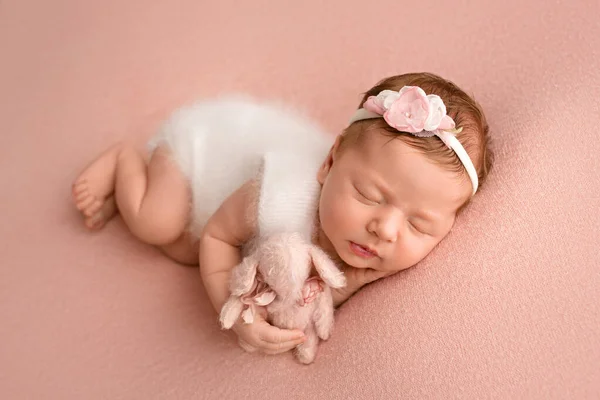ピンクのウサギと白い包帯とピンクの背景に彼女の頭の上にピンクの花を持つ白いジャンプスーツで寝て新生児の女の子のトップビュー 女の子の美しい肖像画7日 1週間 — ストック写真