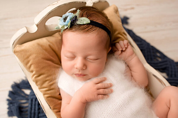 A cute newborn baby girl in a white jumpsuit with a flower bandage sleeps in a wooden crib on a brown pillow, top view. On a light wooden background