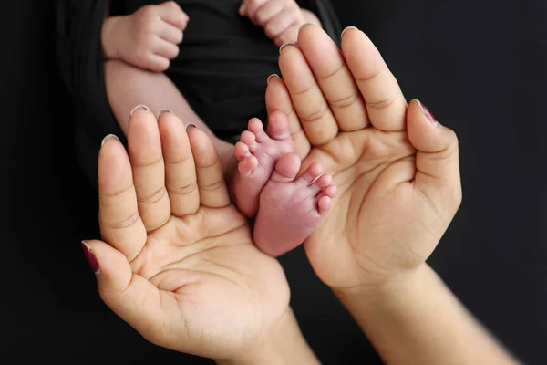 The palms of the parents. A mother hold the feet of a newborn child in a black blanket on a Black background. The feet of a newborn in the hands of parents. Photo of foot, heels and toes.
